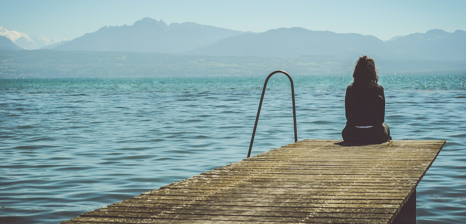 a woman sits on the end of a dock during daytime staring across a lake